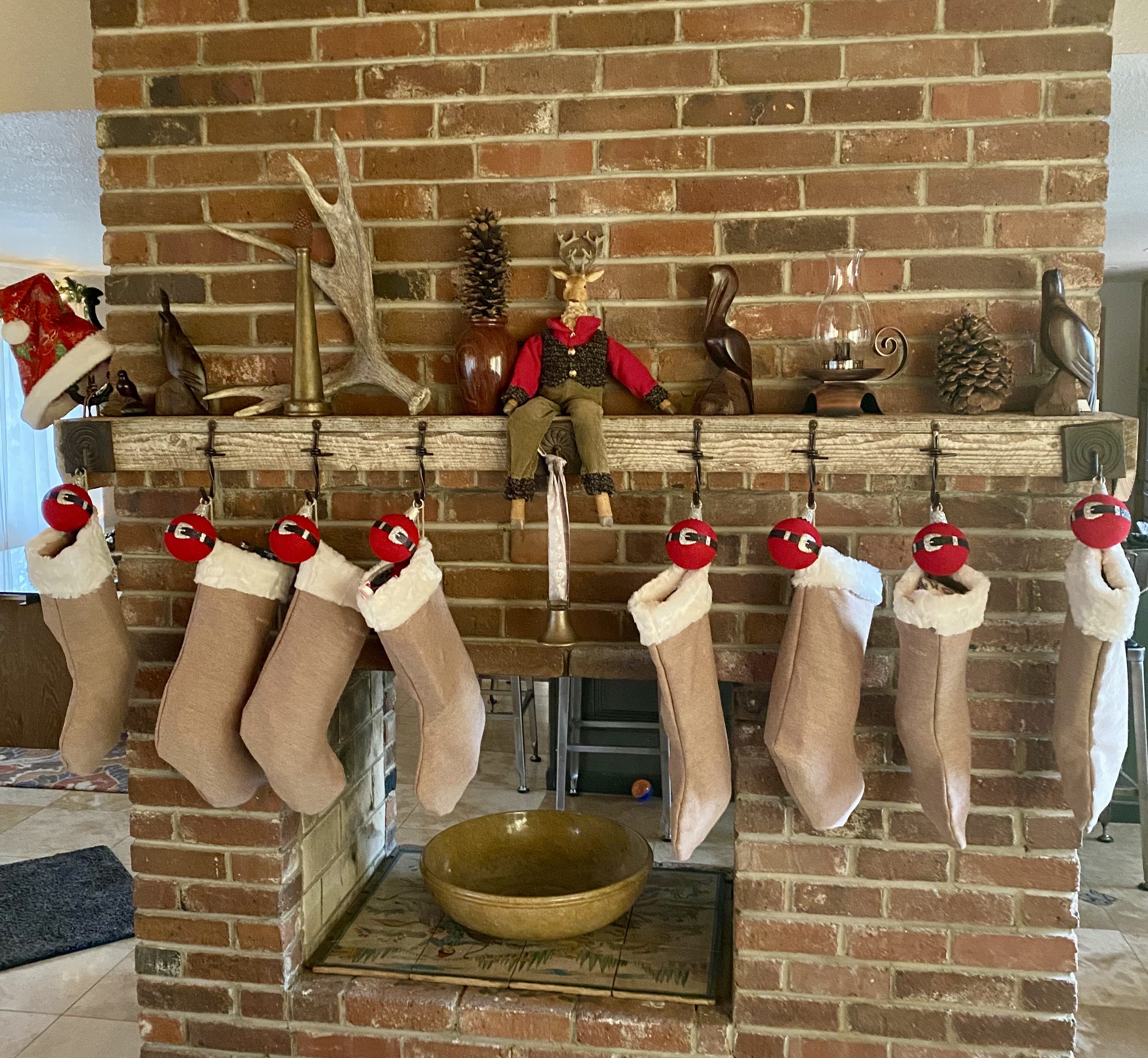 Nine Christmas stockings hanging from a brick fireplace mantel decorated with collected holiday pieces.
